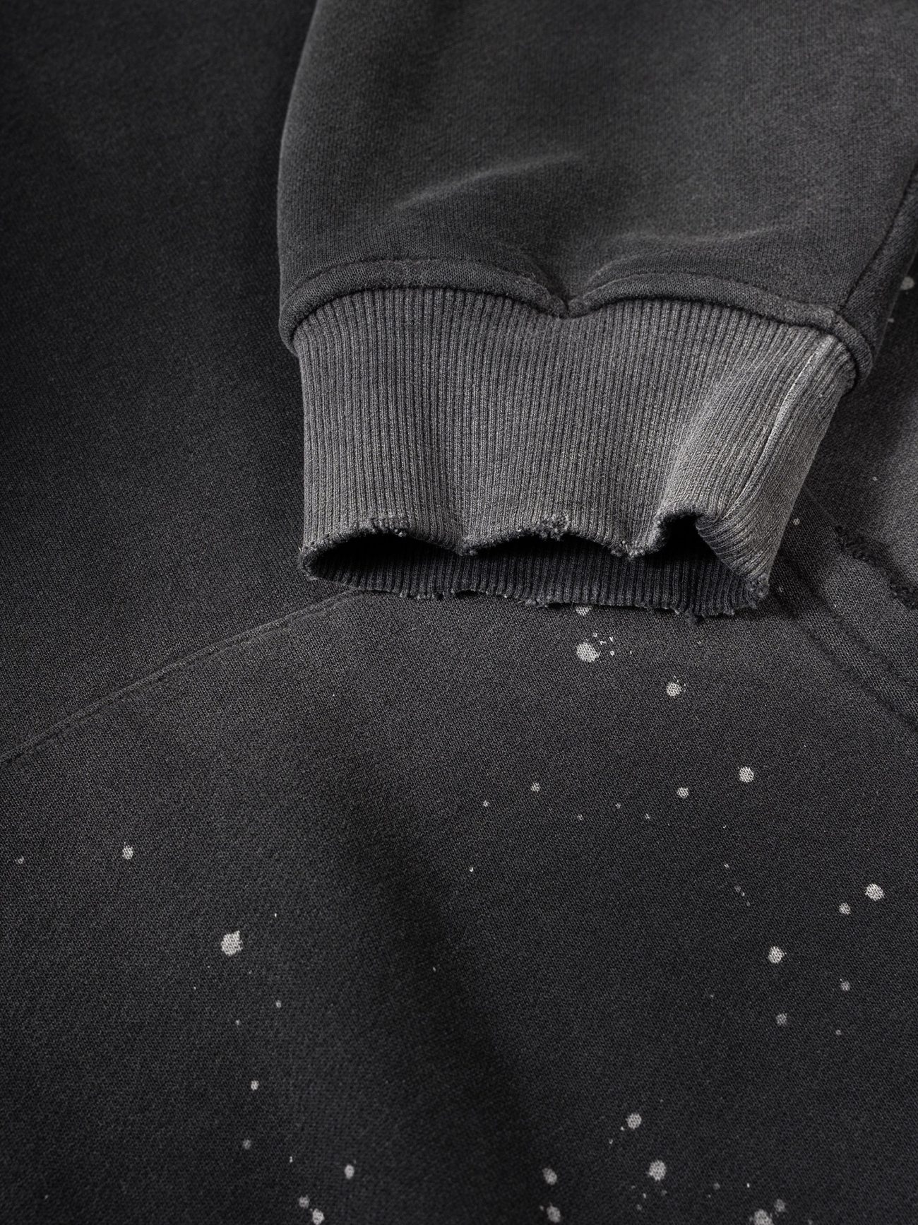A close-up of the Fusion Core - Vintage Wash Frayed Fleece Hoodie (360 GSM) shows a black sleeve with a frayed cuff and white paint splatters, highlighting the edgy details of this affordable luxury piece.
