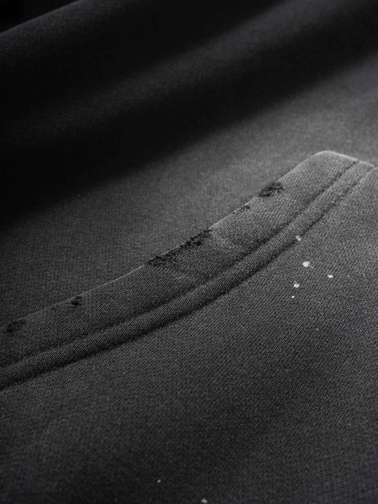 Close-up of the Fusion Core - Vintage Wash Frayed Fleece Hoodie (360 GSM) in black, featuring a seam and pocket with white paint splatters and frayed edges—details that showcase its affordable luxury appeal.