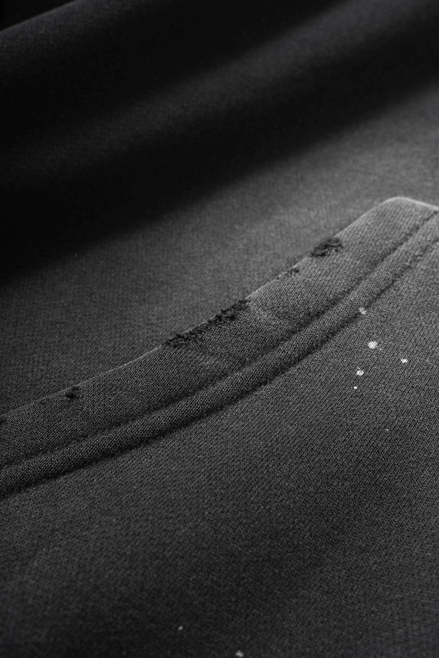 Close-up of the Fusion Core - Vintage Wash Frayed Fleece Hoodie (360 GSM) in black, featuring a seam and pocket with white paint splatters and frayed edges—details that showcase its affordable luxury appeal.