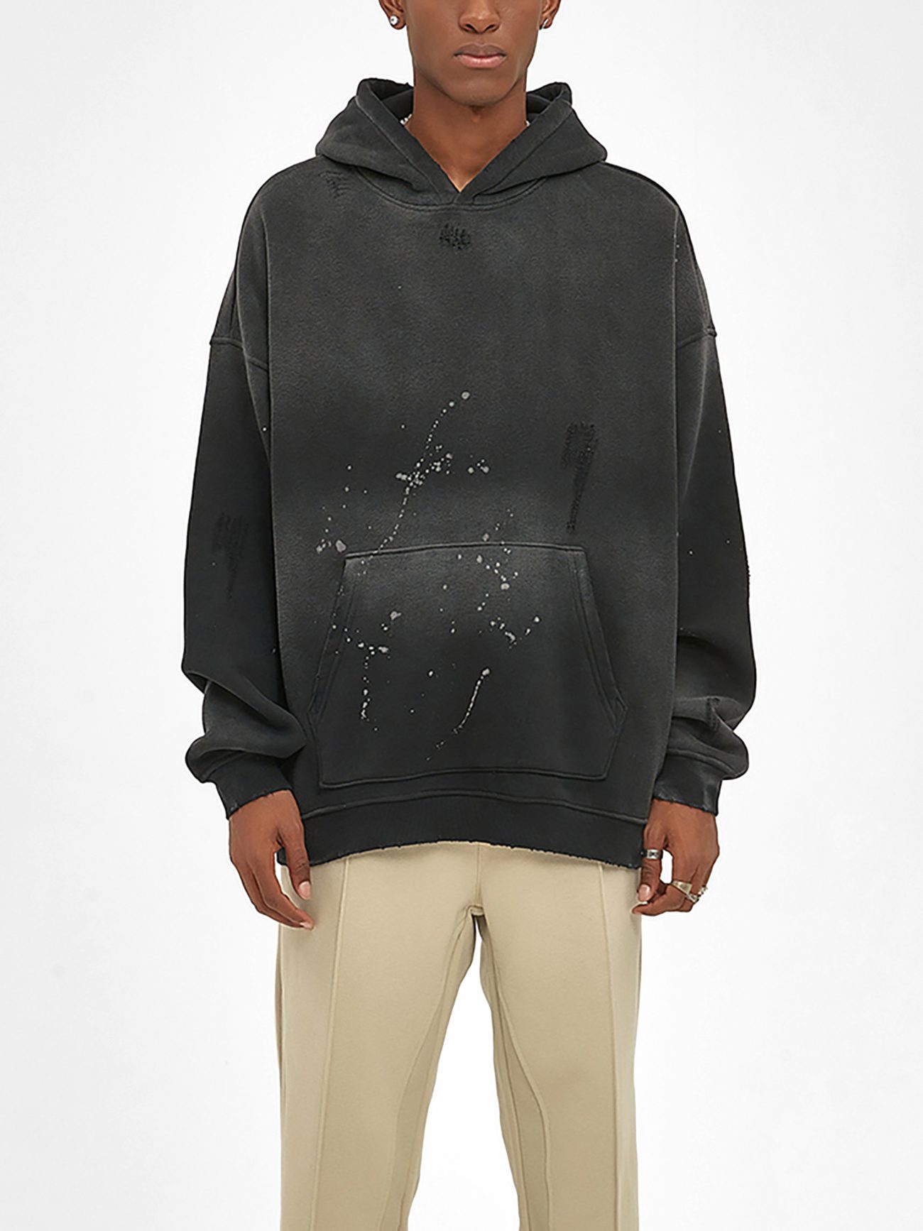 A person wears the Fusion Core - Vintage Wash Frayed Fleece Hoodie (360 GSM), black and oversized with paint splatter details, front pocket, and hood, paired with beige pants to showcase Affordable Luxury Clothing against a plain white background.