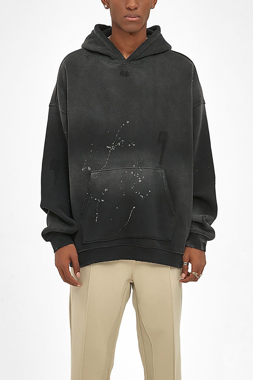 A person wears the Fusion Core - Vintage Wash Frayed Fleece Hoodie (360 GSM), black and oversized with paint splatter details, front pocket, and hood, paired with beige pants to showcase Affordable Luxury Clothing against a plain white background.