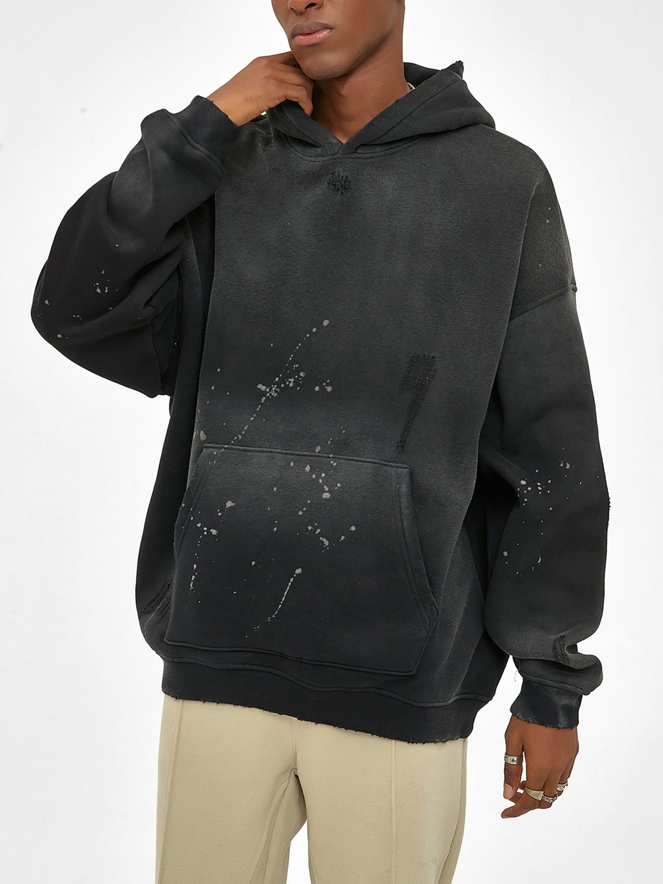 Wearing the Fusion Core - Vintage Wash Frayed Fleece Hoodie (360 GSM), a person in beige pants stands against a white background. The hoodie features paint splatters, blending affordable luxury with a relaxed fit; their hand touches their chin, face partly out of frame.