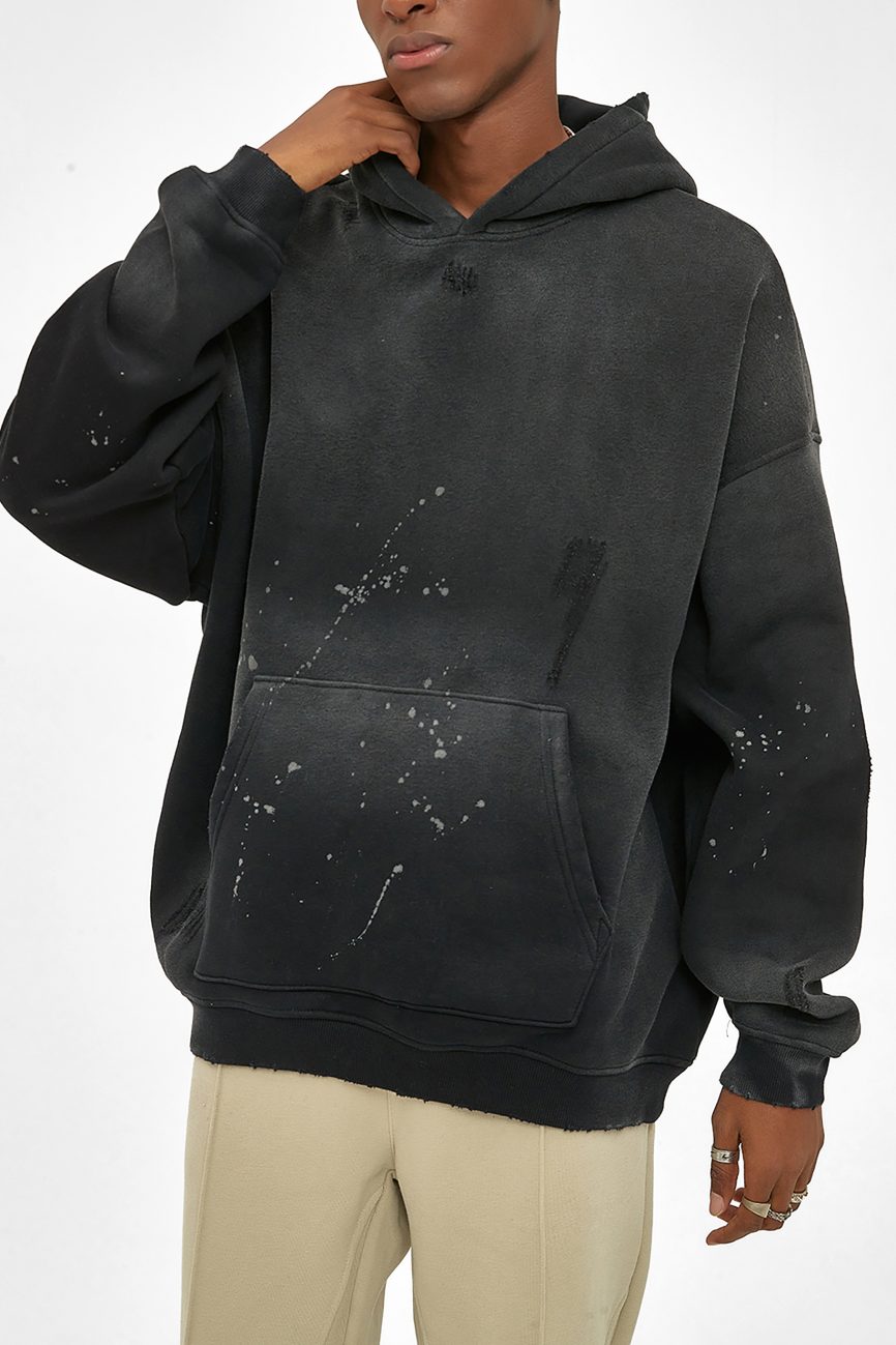 Wearing the Fusion Core - Vintage Wash Frayed Fleece Hoodie (360 GSM), a person in beige pants stands against a white background. The hoodie features paint splatters, blending affordable luxury with a relaxed fit; their hand touches their chin, face partly out of frame.