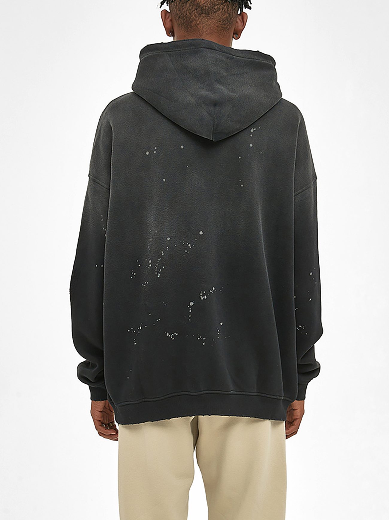 A person stands with their back to the camera, wearing the Fusion Core Vintage Wash Frayed Fleece Hoodie (360 GSM) in black with paint splatters and light beige pants, set against a plain white background.