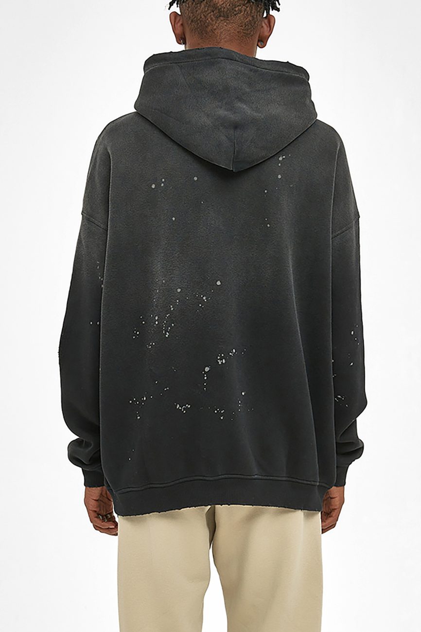 A person stands with their back to the camera, wearing the Fusion Core Vintage Wash Frayed Fleece Hoodie (360 GSM) in black with paint splatters and light beige pants, set against a plain white background.