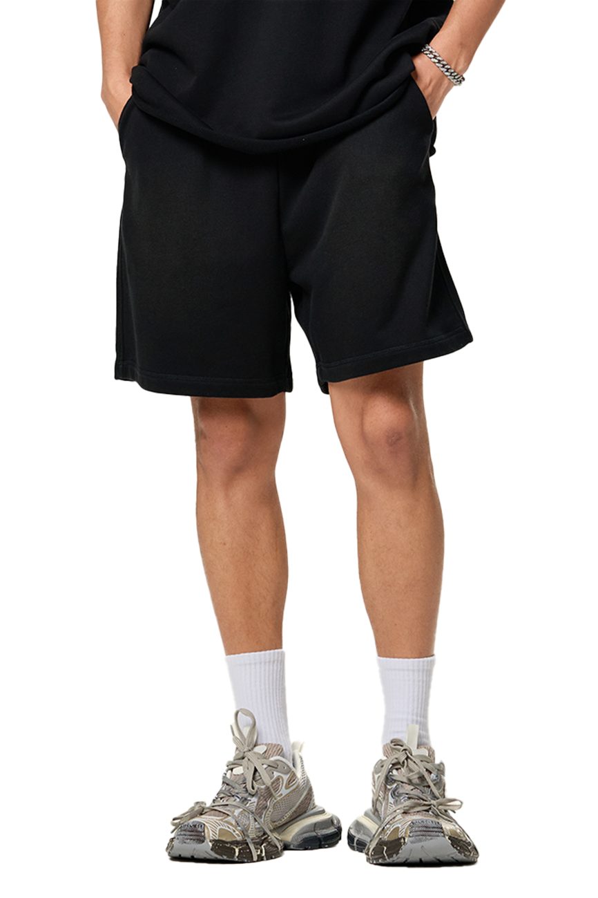 A person wearing affordable luxury stands with hands in pockets, shown from the waist down in Sun Fade Raw Edge Cotton Shorts paired with a black t-shirt, white socks, and gray sneakers.