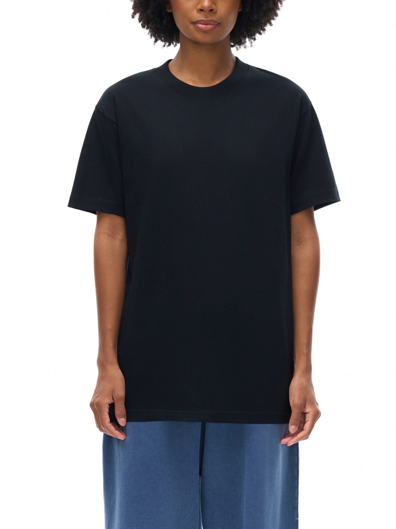 A person with short curly hair wears a Fusion Emblem - Oversized T-shirt in plain black and loose blue pants, standing against a white background. Their face is partially visible.