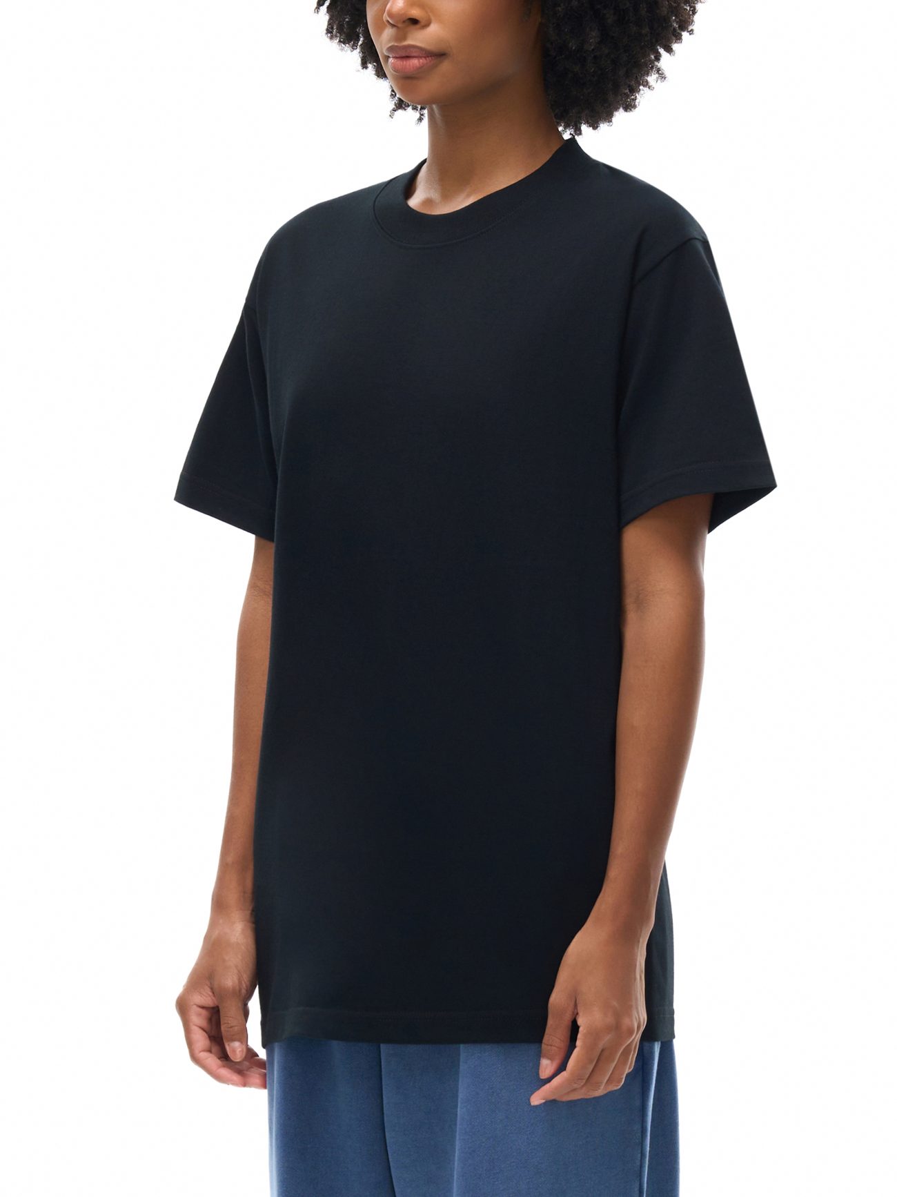A person with curly hair models the Fusion Emblem - Oversized T-shirt in plain black, paired with blue pants—an effortless display of affordable luxury—against a white background. Only their upper body is visible.