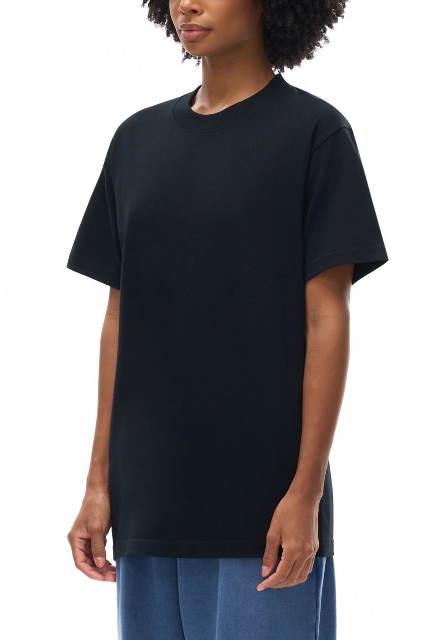 A person with curly hair models the Fusion Emblem - Oversized T-shirt in plain black, paired with blue pants—an effortless display of affordable luxury—against a white background. Only their upper body is visible.