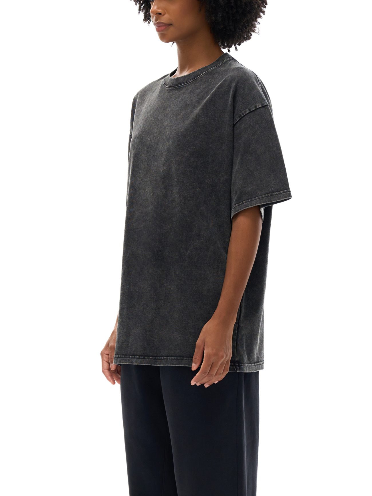 A person stands in profile wearing the Acid Wash Oversize T-Shirt - 250 GSM, styled effortlessly with dark pants. Their curly hair and relaxed pose complete the look against a white background, capturing modern, understated style.