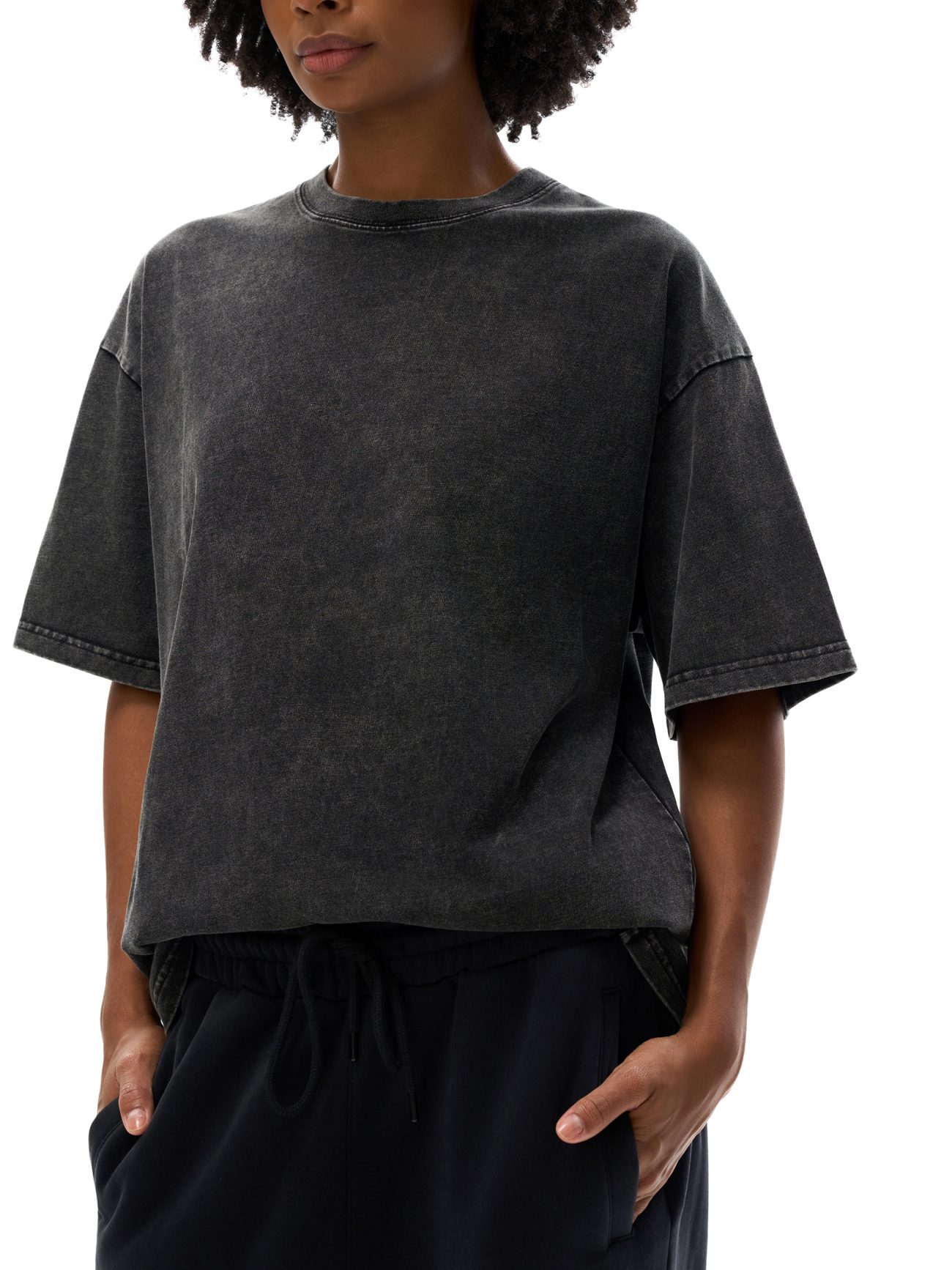 A woman stands against a plain white background, hands in her pockets, wearing the Fifteen Acid Wash Oversize T-Shirt (250 GSM) in faded black and black pants—an effortless take on affordable luxury style.