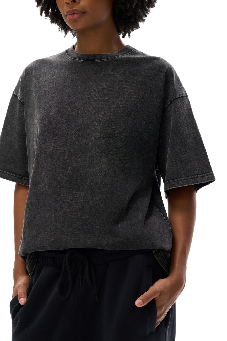 A woman stands against a plain white background, hands in her pockets, wearing the Fifteen Acid Wash Oversize T-Shirt (250 GSM) in faded black and black pants—an effortless take on affordable luxury style.