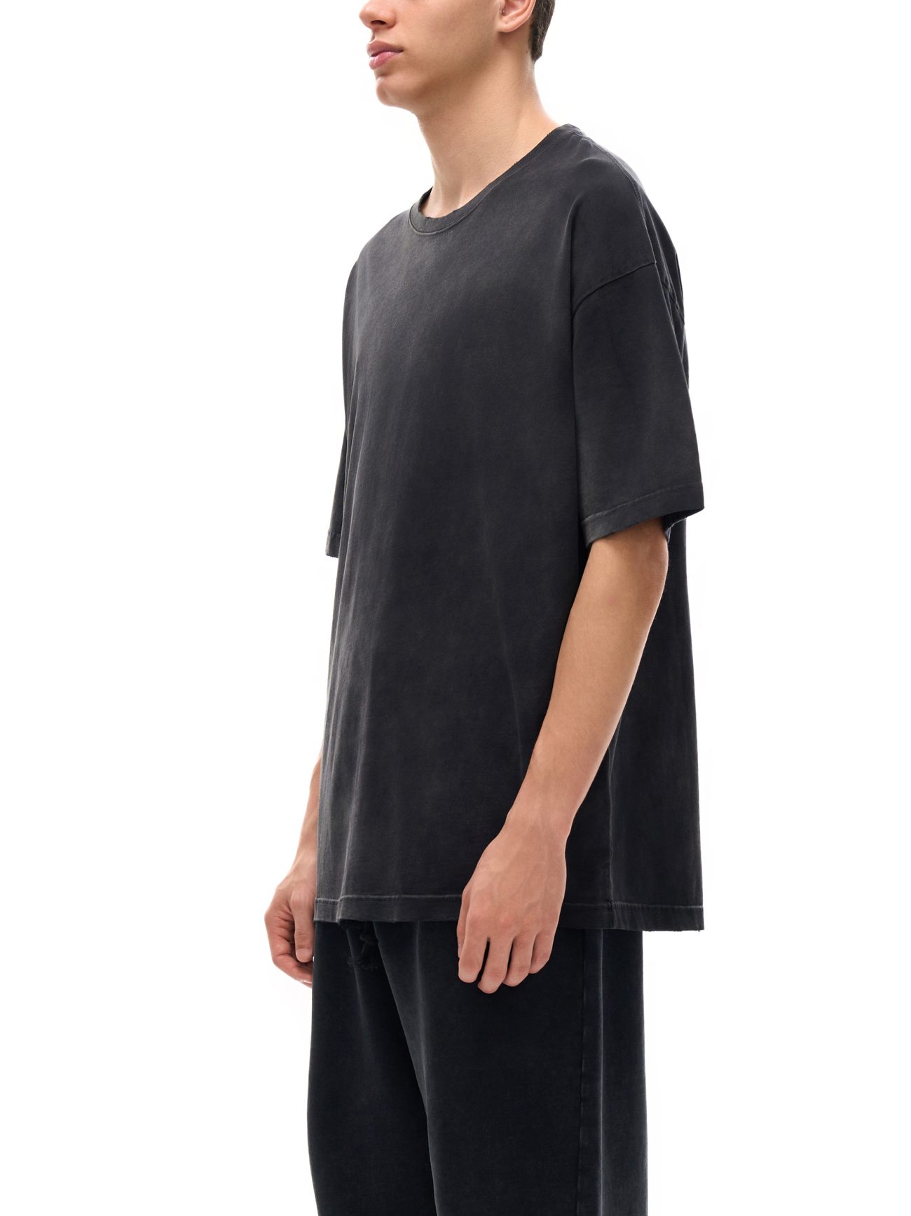 Wearing the Fusion Atom - Vintage Washed Hand-Frayed T-Shirt, a person in loose-fitting faded black attire stands sideways against a white background, their face partially out of view—embodying affordable luxury style.
