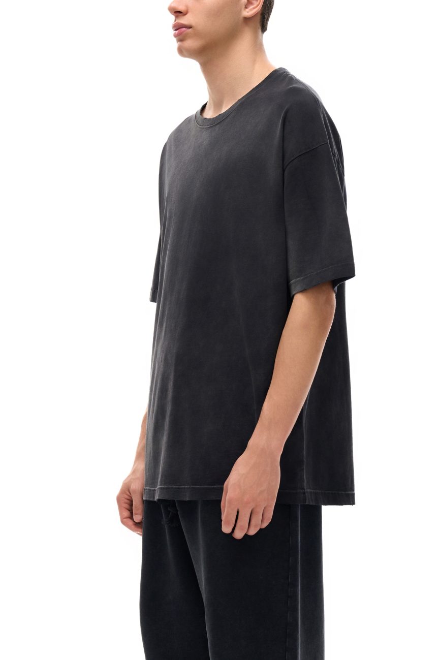 Wearing the Fusion Atom - Vintage Washed Hand-Frayed T-Shirt, a person in loose-fitting faded black attire stands sideways against a white background, their face partially out of view—embodying affordable luxury style.