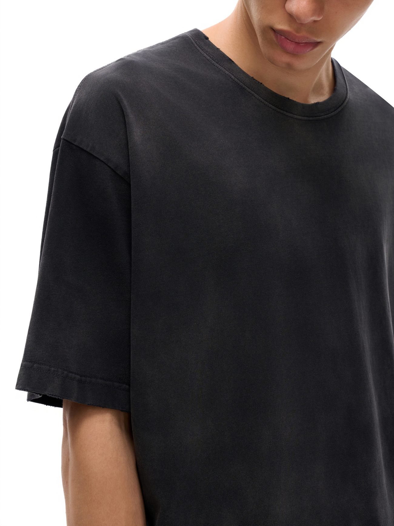 A person wears the Fusion Atom - Vintage Washed Hand-Frayed T-Shirt, a loose-fitting faded black tee from an affordable luxury brand, shown from shoulders to upper chest against a white background, face partially visible and looking down.