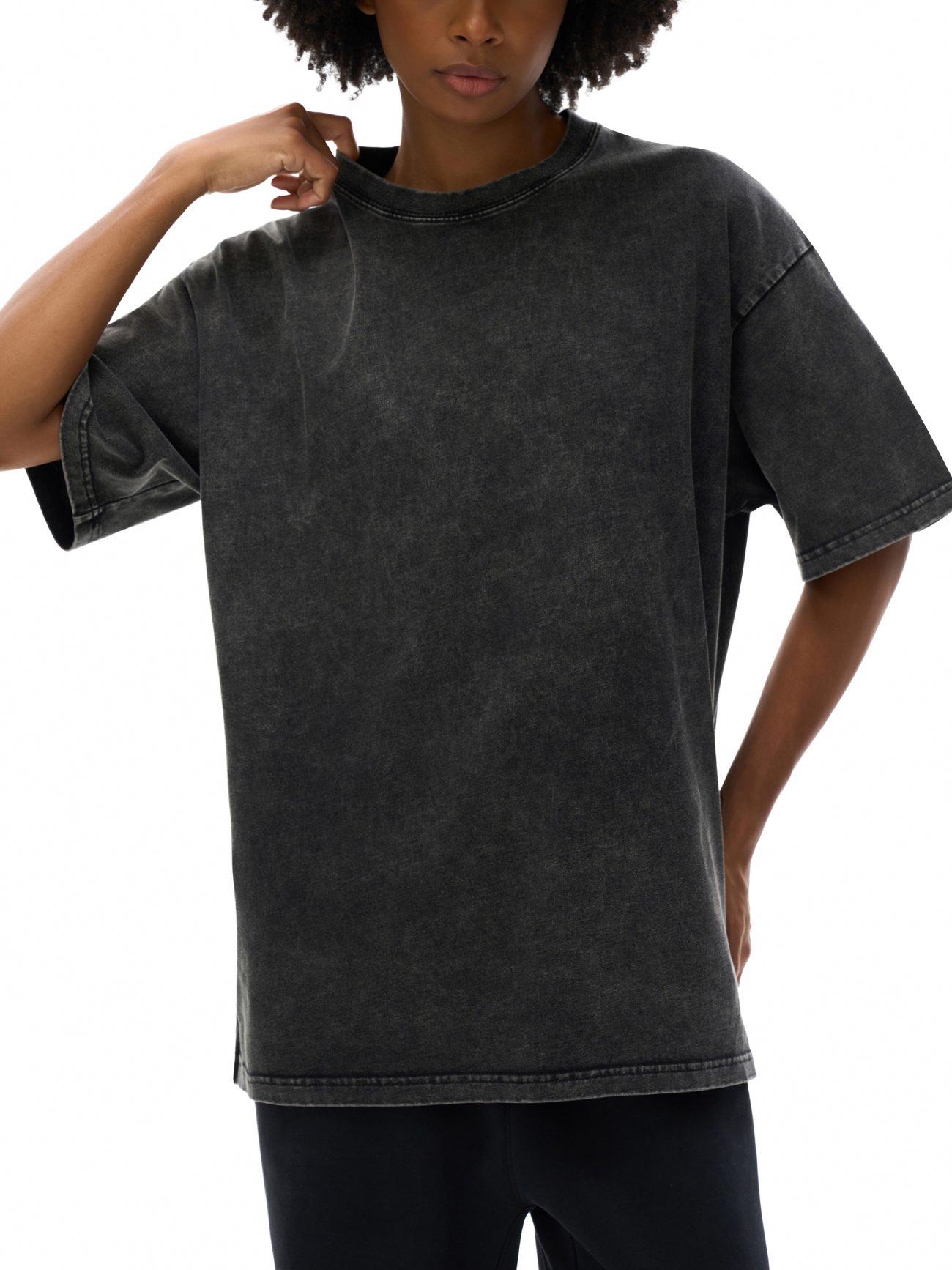 A person in the Fifteen Acid Wash Oversize T-Shirt (250 GSM)—a loose-fitting, washed black tee with short sleeves—and dark pants stands against a white background, lightly tugging at the collar.