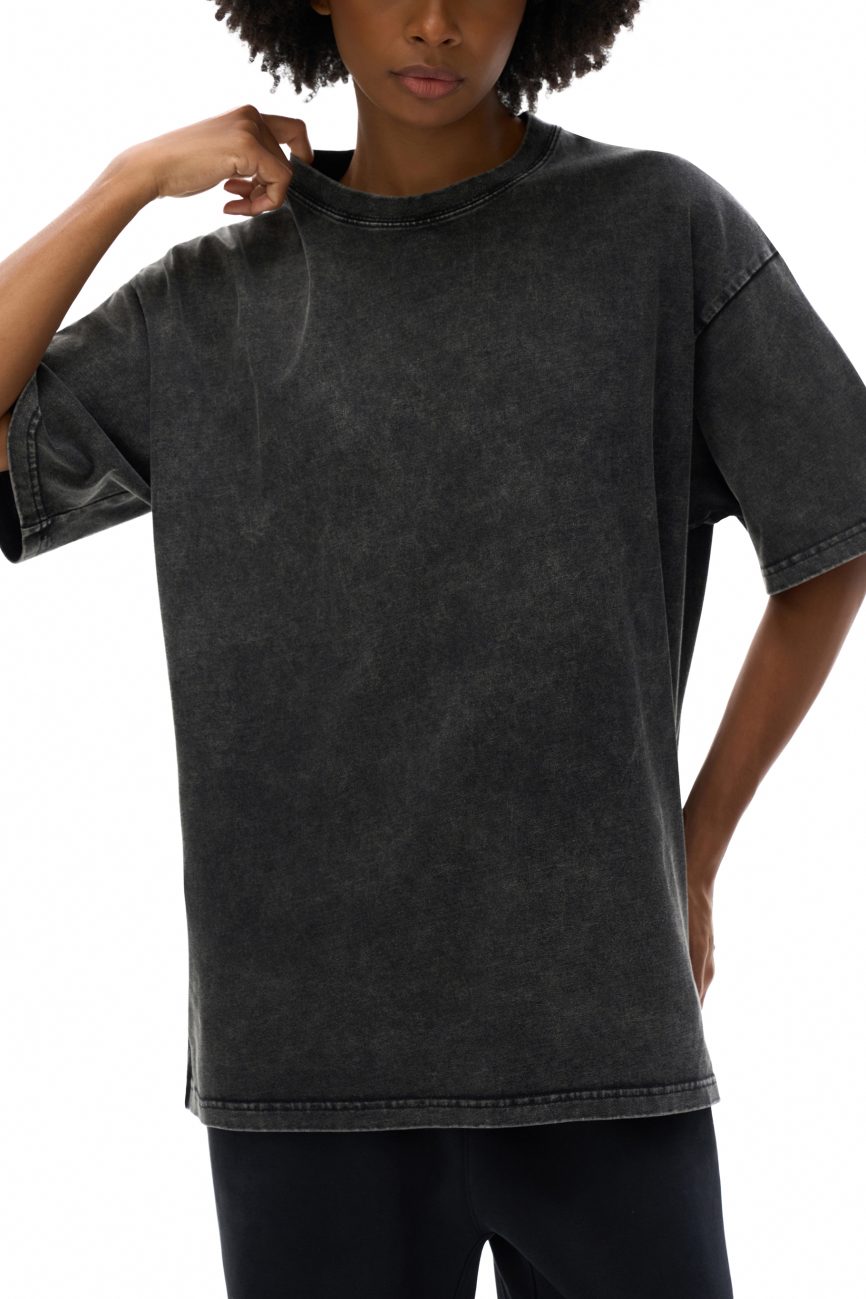 A person in the Fifteen Acid Wash Oversize T-Shirt (250 GSM)—a loose-fitting, washed black tee with short sleeves—and dark pants stands against a white background, lightly tugging at the collar.