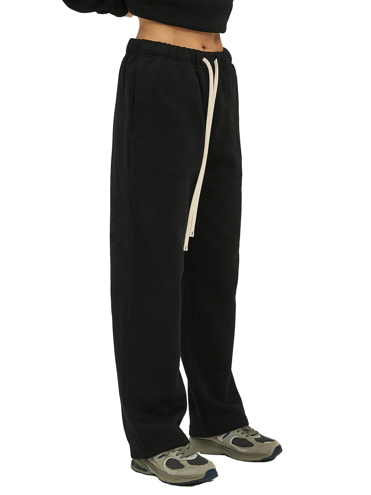 A person wears Fusion Streetwear Unisex Solid Color Fleece Straight Leg Pants in black, featuring a long white drawstring, paired with green and gray sneakers. Only the lower torso and legs are shown against a white background.