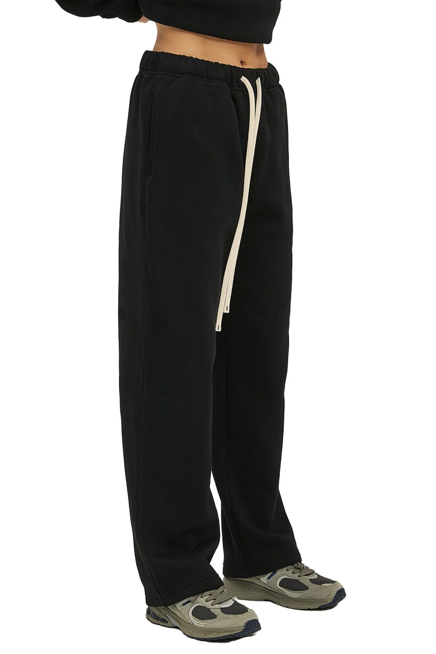 A person wears Fusion Streetwear Unisex Solid Color Fleece Straight Leg Pants in black, featuring a long white drawstring, paired with green and gray sneakers. Only the lower torso and legs are shown against a white background.