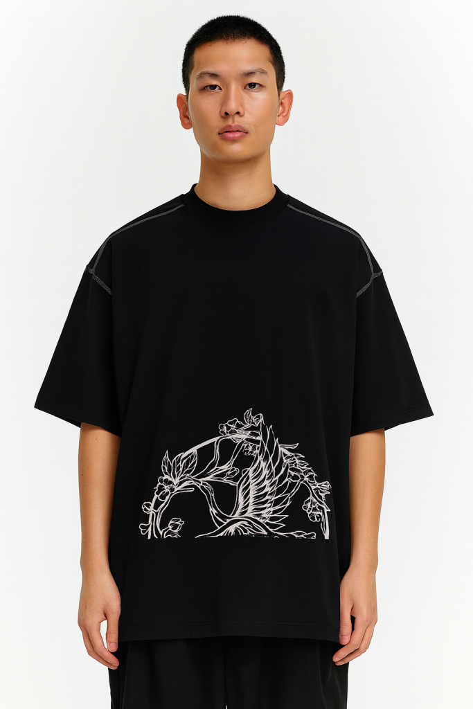 A person faces forward, wearing the Crane - Oversized Contrast-Stitched Cotton Tee, an affordable luxury black T-shirt with a white outlined bird and floral design on the lower front. The background is plain white.