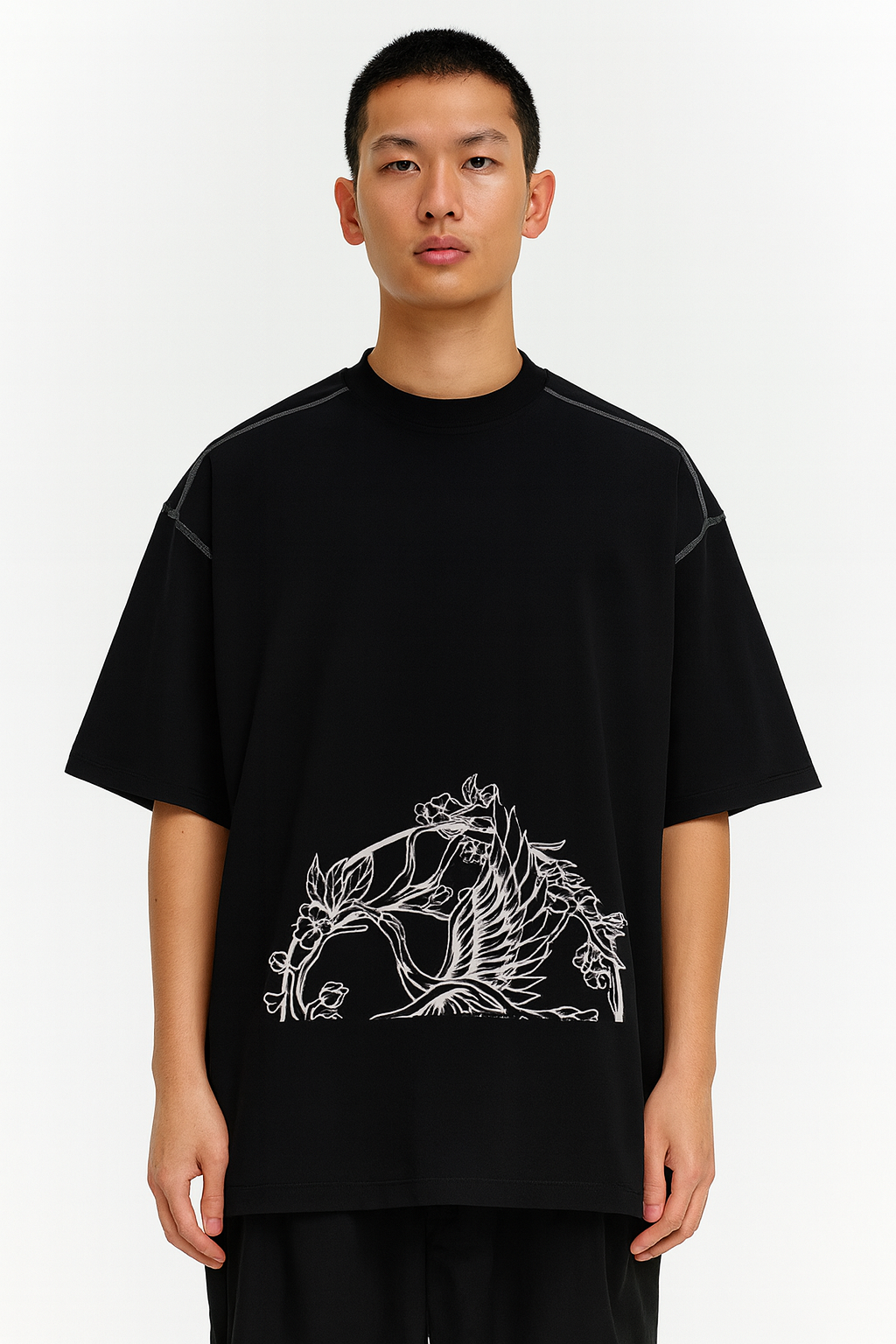 A person faces forward, wearing the Crane - Oversized Contrast-Stitched Cotton Tee, an affordable luxury black T-shirt with a white outlined bird and floral design on the lower front. The background is plain white.
