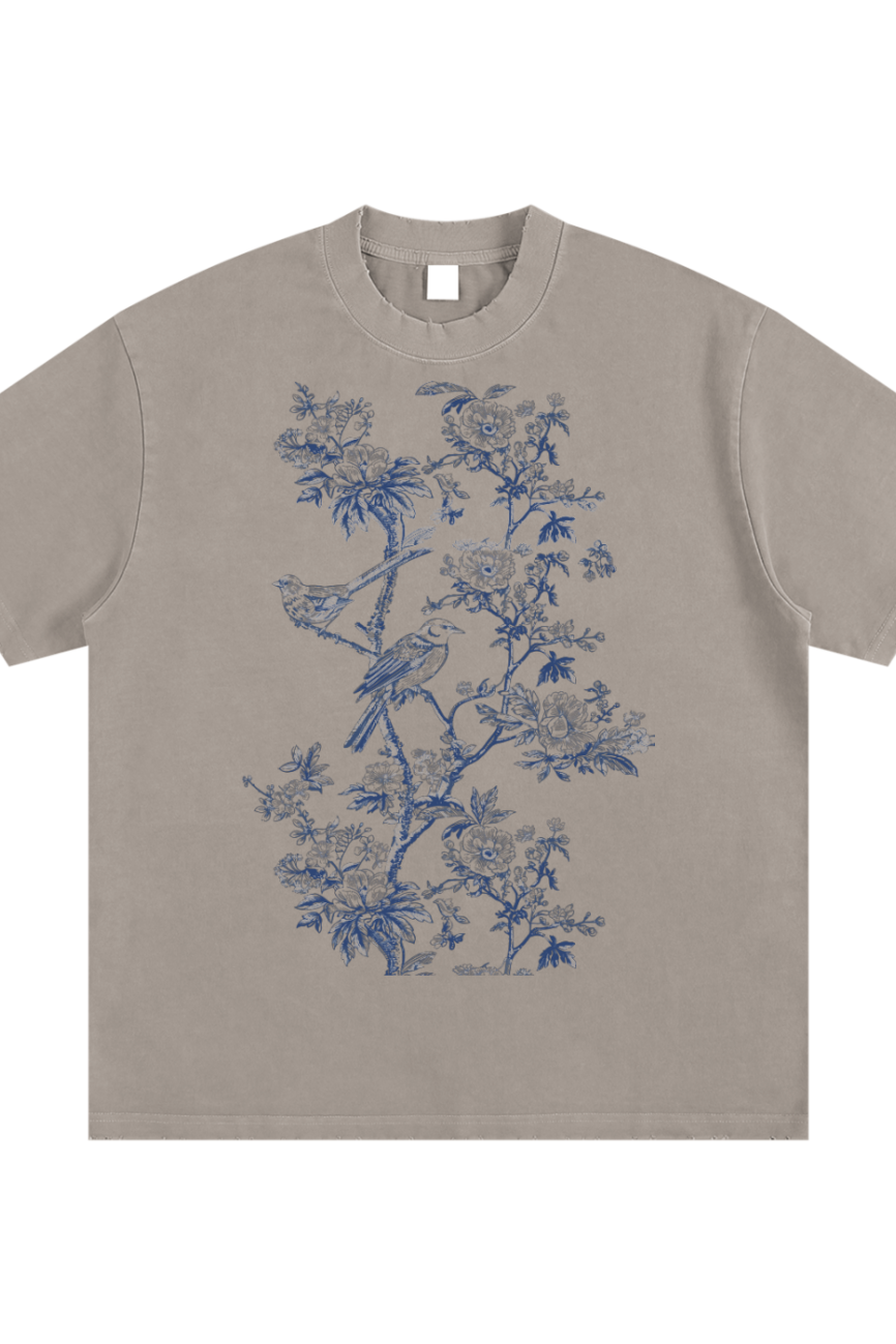 The Toile De Jouy Heavyweight Vintage Washed Frayed T-Shirt is a taupe short-sleeve tee with a detailed blue floral and bird print on the front center, offering affordable luxury for your everyday style.