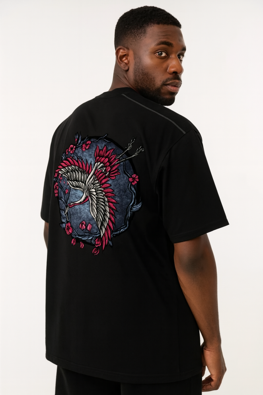 A man wears the Crane - Oversized Contrast-Stitched Cotton Tee, a black affordable luxury shirt with a vibrant embroidered crane and red flowers on the back, as he looks over his shoulder against a plain light background.