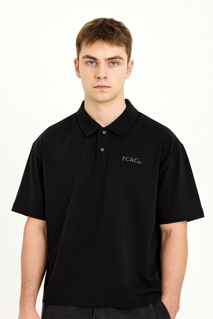 A young man with short brown hair models the FC&Co. Drop Shoulder Short-Sleeve Polo, a loose-fitting black shirt with an embroidered logo, representing affordable luxury against a plain white background.