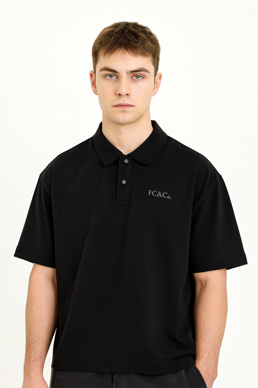 A young man with short brown hair models the FC&Co. Drop Shoulder Short-Sleeve Polo, a loose-fitting black shirt with an embroidered logo, representing affordable luxury against a plain white background.
