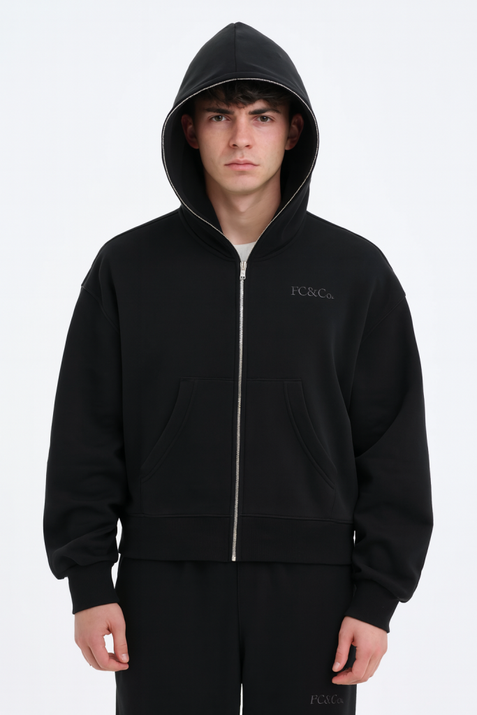 A young man wears the FC&Co. Fleeced Full-Zip Boxy Hoodie with the hood up, featuring a small chest logo. He pairs it with matching black pants and stands against a plain white background. Part of our Affordable Luxury Clothing line.