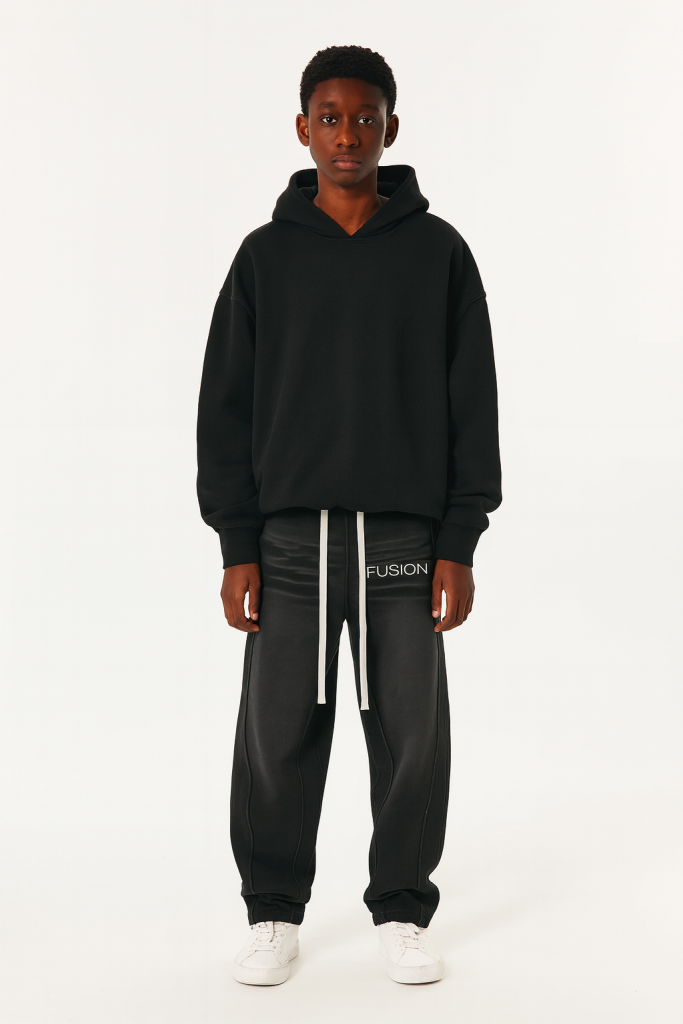 A young person faces forward in Sunfade Curved Blade Fleece Sweatpants and a loose black hoodie—affordable luxury attire—paired with white sneakers, all set against a plain white background.
