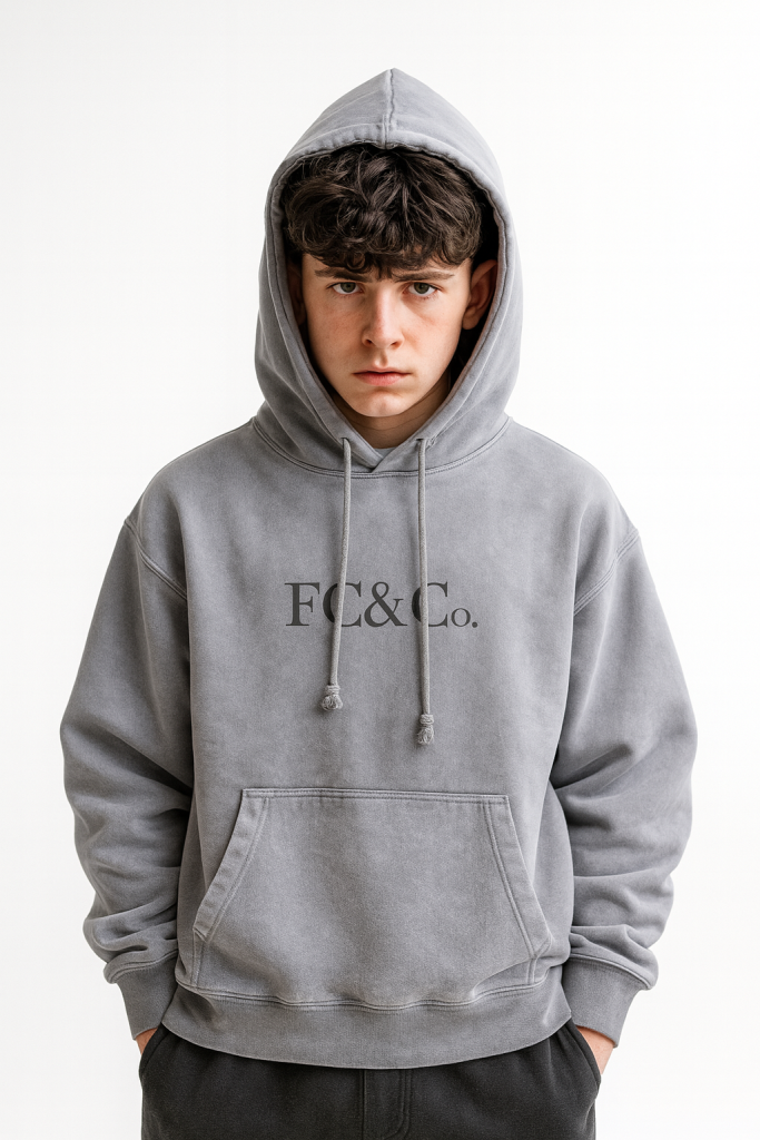 A young person with short, curly dark hair wears the FC&Co. Ombre Vintage Stone Washed Colorpointed Hoodie, standing against a plain white background with hands in the pocket and looking directly at the camera—an effortless, affordable luxury look.