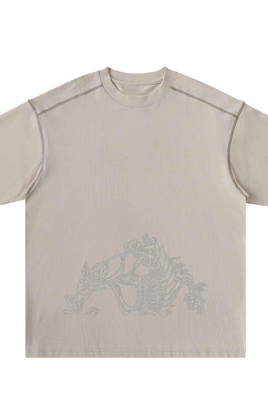 The Crane - Oversized Contrast-Stitched Cotton Tee features a beige, short-sleeve design with subtle light gray floral line art near the bottom front and visible shoulder seam stitching for an affordable touch of luxury.