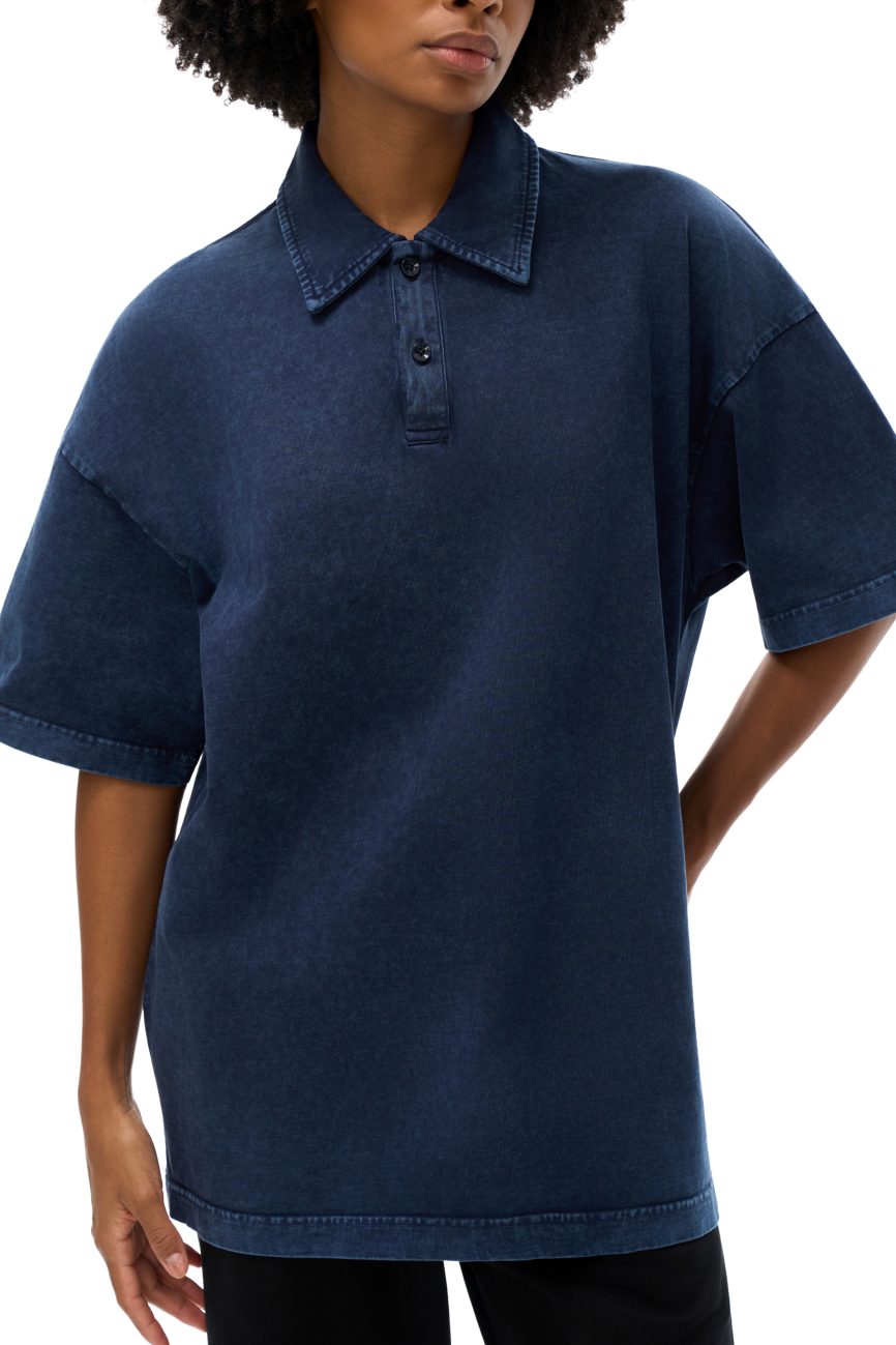 A person wearing the Fusion Heavyweight Snow-Washed Polo T-Shirt—a loose-fitting, short-sleeved, dark blue denim polo that represents affordable luxury—stands confidently with one hand on their hip.
