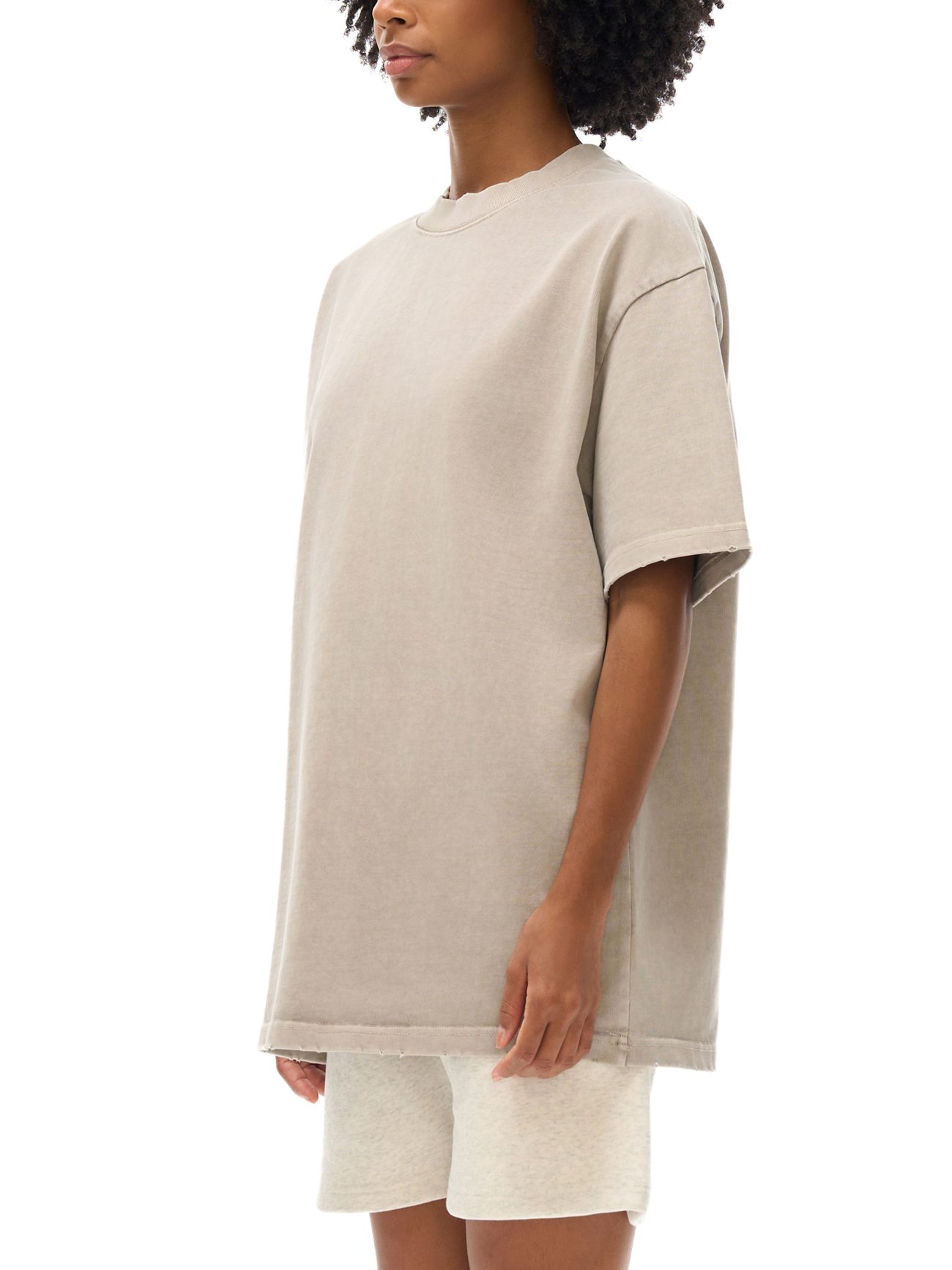 A person with curly hair models the Toile De Jouy Heavyweight Vintage Washed Frayed T-Shirt in light beige, paired with matching shorts. The relaxed-fit outfit is shown against a white background, with the face partially out of the frame.