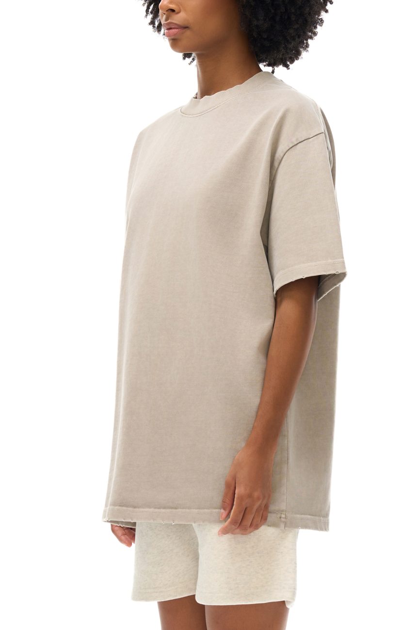 A person with curly hair models the Toile De Jouy Heavyweight Vintage Washed Frayed T-Shirt in light beige, paired with matching shorts. The relaxed-fit outfit is shown against a white background, with the face partially out of the frame.