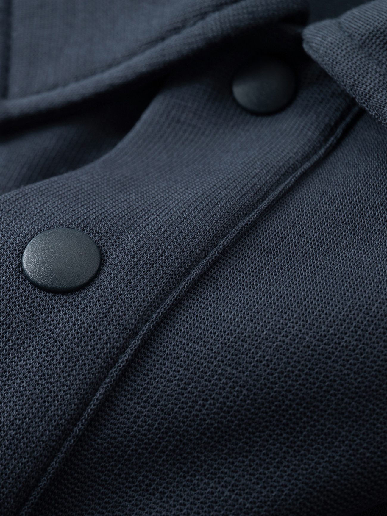 Close-up of the FC&Co. Drop Shoulder Short-Sleeve Polo shows dark textured fabric and two black snap buttons, highlighting the detailed weave and stitching—an example of affordable luxury with attention to detail.