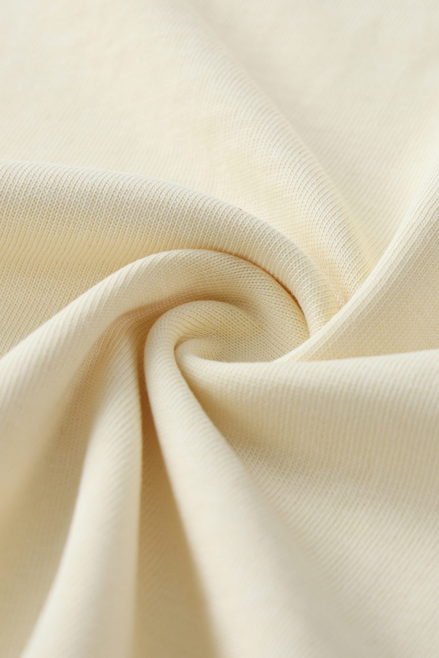 A close-up of the Crane Oversized Contrast-Stitched Cotton Tee shows cream cotton fabric with a smooth, soft texture in a twisted fold, highlighting its weave and gentle drape—ideal for affordable luxury clothing.